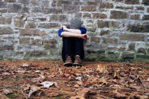 un adolescent adossé contre un mur de brique en extérieur, il est assi au sol, il enlace ses genoux qui son joint contre son visage. Il y a des feuille au sol